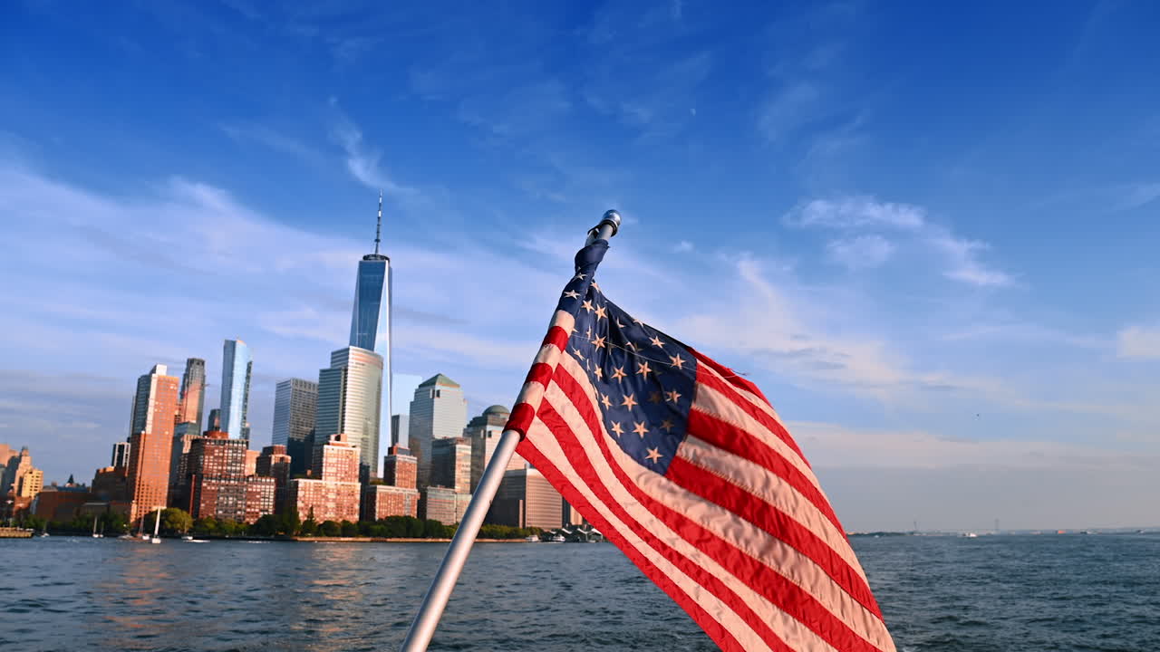 Waving American flag in the light of bright sun. Skyline of New York at backdrop. View from the river