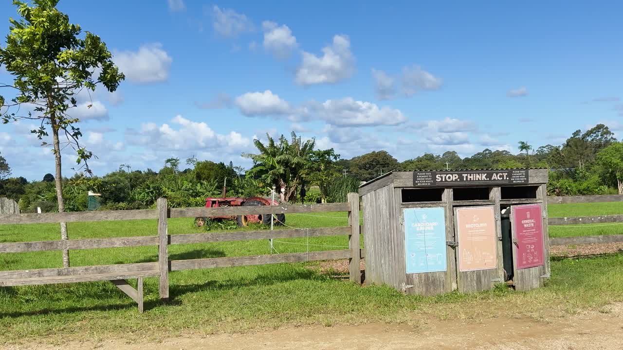 Three waste separation bins stand near a wooden fence in a grassy rural field under bright daylight, with static camera and clear blue sky