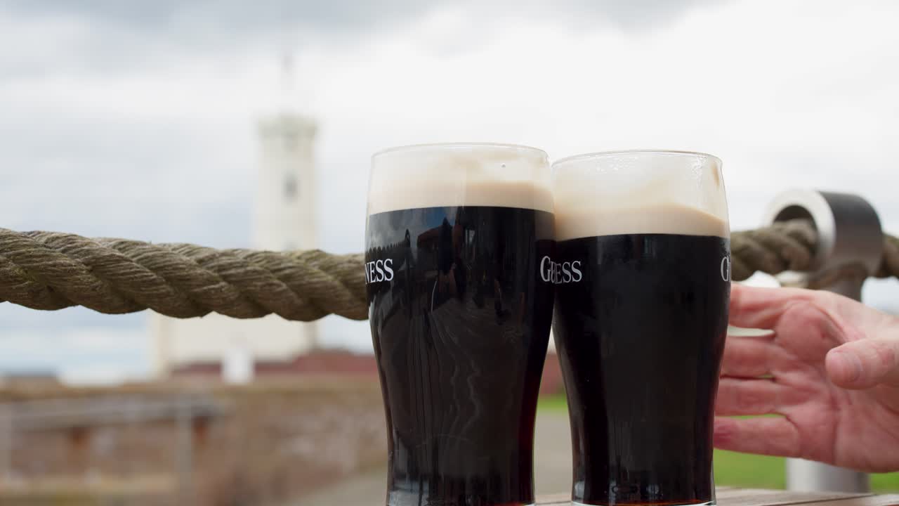 Two hands clink dark beer glasses outdoors, overcast daylight, shallow depth of field, steady shot