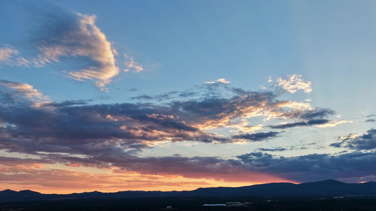 A drone timelapse of a sunset ray of orange light moving and fading over a southwest mountain horizon, illuminating the scattered cloud layers above the dark silhouette below.