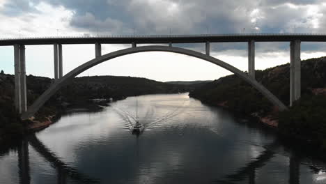 Barco-De-Vela-Siguiendo-La-Cámara-Bajo-Un-Puente-De-Coche-En-Un-Fiordo-En-Croacia