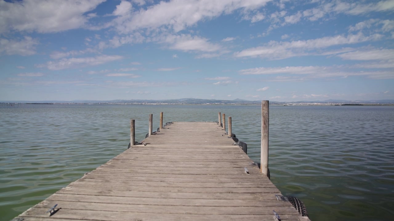 Plano de seguimiento en primera persona (POV) moviéndose a través de una puerta hacia un muelle de madera que da a un lago tranquilo con montañas, día de verano, cámara lenta