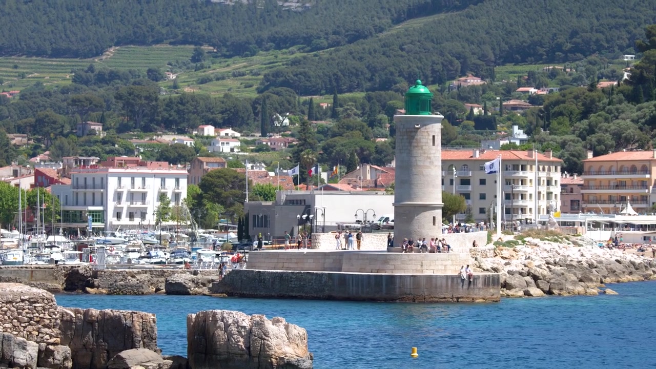 Inquadratura ampia di un faro in pietra e di un porto con montagne sotto la luce intensa del giorno.