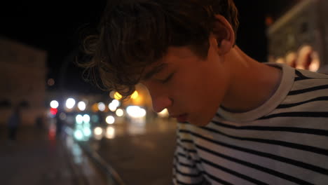 solitary teenage male standing beside urban street, experiencing emotional moment with blurred car lights and architectural background during nighttime cityscape
