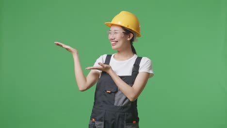 asian woman worker wearing goggles and safety helmet smiling and pointing to side while standing in the green screen background studio