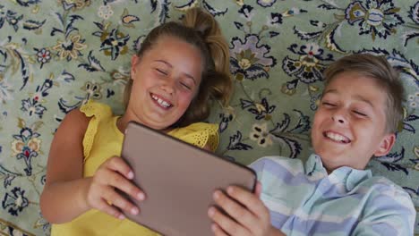 Happy-caucasian-brother-and-sister-at-home,-lying-on-floor-using-tablet-together-and-smiling