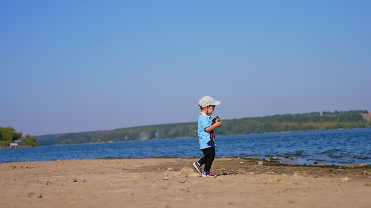 Cute baby boy running cheerfully by the river bank. Kid picks the stone and carries it to throw into water.