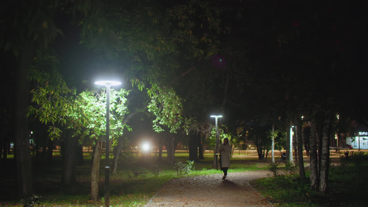 Young adult walking along peaceful night path with lush trees and glowing lampstands, creating a calm serene ambiance while distant car lights softly illuminate background in quiet urban park setting