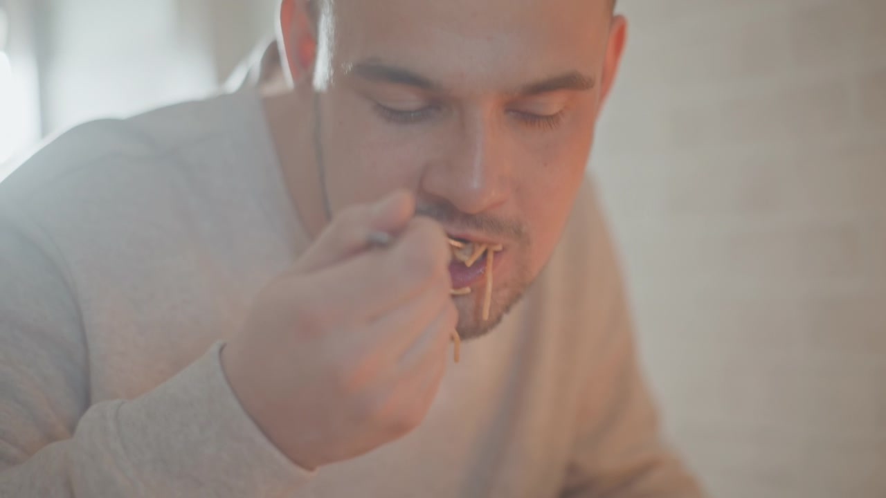 Uomo seduto mangia spaghetti da una ciotola bianca, sollevando la forchetta, avvolgendo gli spaghetti, poi abbassando l'utensile mentre mastica; espressione rilassata, luce calda domestica e uno sfondo sfocato creano un'atmosfera accogliente per la cena.