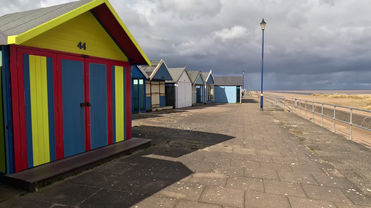 Colorful beach huts stood in a line along the seafront with sandy beach and moody grey sky&rsquo;s