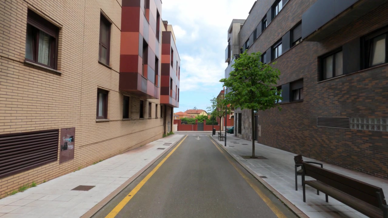 Vista de rua em movimento através de uma tranquila rua residencial ladeada por edifícios de apartamentos modernos de tijolos e árvores em Gijón, Espanha.
