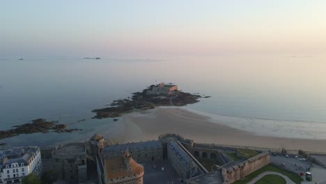 drone shot approaching fort national with saint-malo city, sandbank, and sea at dusk.