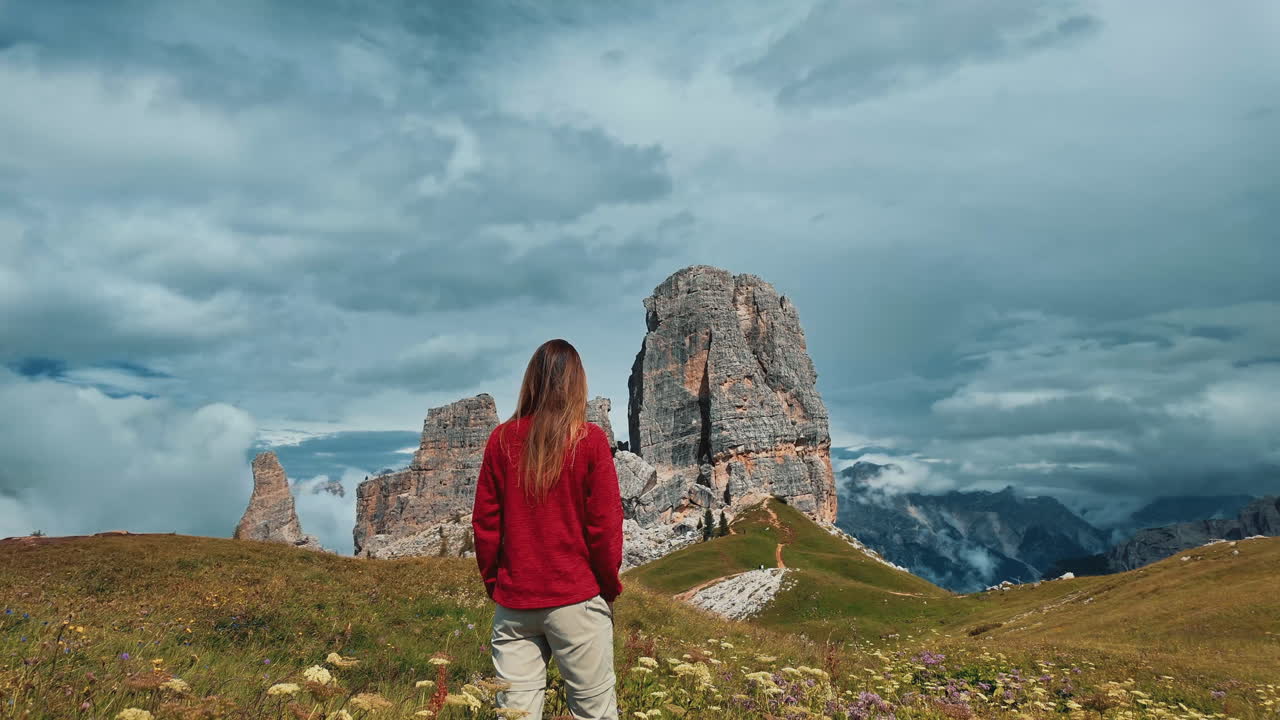 Lone female hiker standing on rocky terrain, gazing at dramatic Cinque Torri rock formations against sweeping mountain landscape in golden summer light