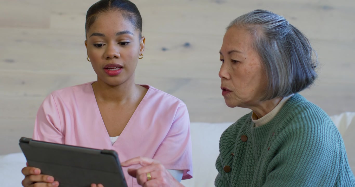 Diverse female caregiver in pink scrubs offering tablet while teaching Senior client at home