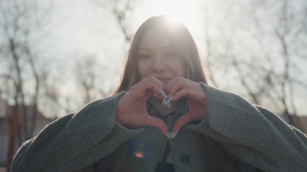 Hübsche Frau im Gegenlicht macht eine Herzgeste mit Sonnenlicht-Halo, Hände bilden ein Herz, freudiges Lächeln, Wintermantel, Stadtpark, liebevolle Stimmung, Lens Flare, authentisches Porträt, zärtlicher Ausdruck, filmische Wärme.