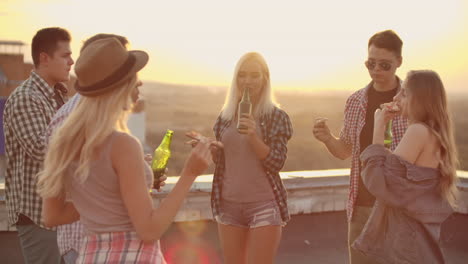 The-company-of-six-young-people-enjoy-the-party-on-the-roof.-They-eat-hot-pizza-and-drink-beer-from-the-green-bottles-they-smiles-on-a-summer-evening.-They-dance-and-communicate-with-each-other.