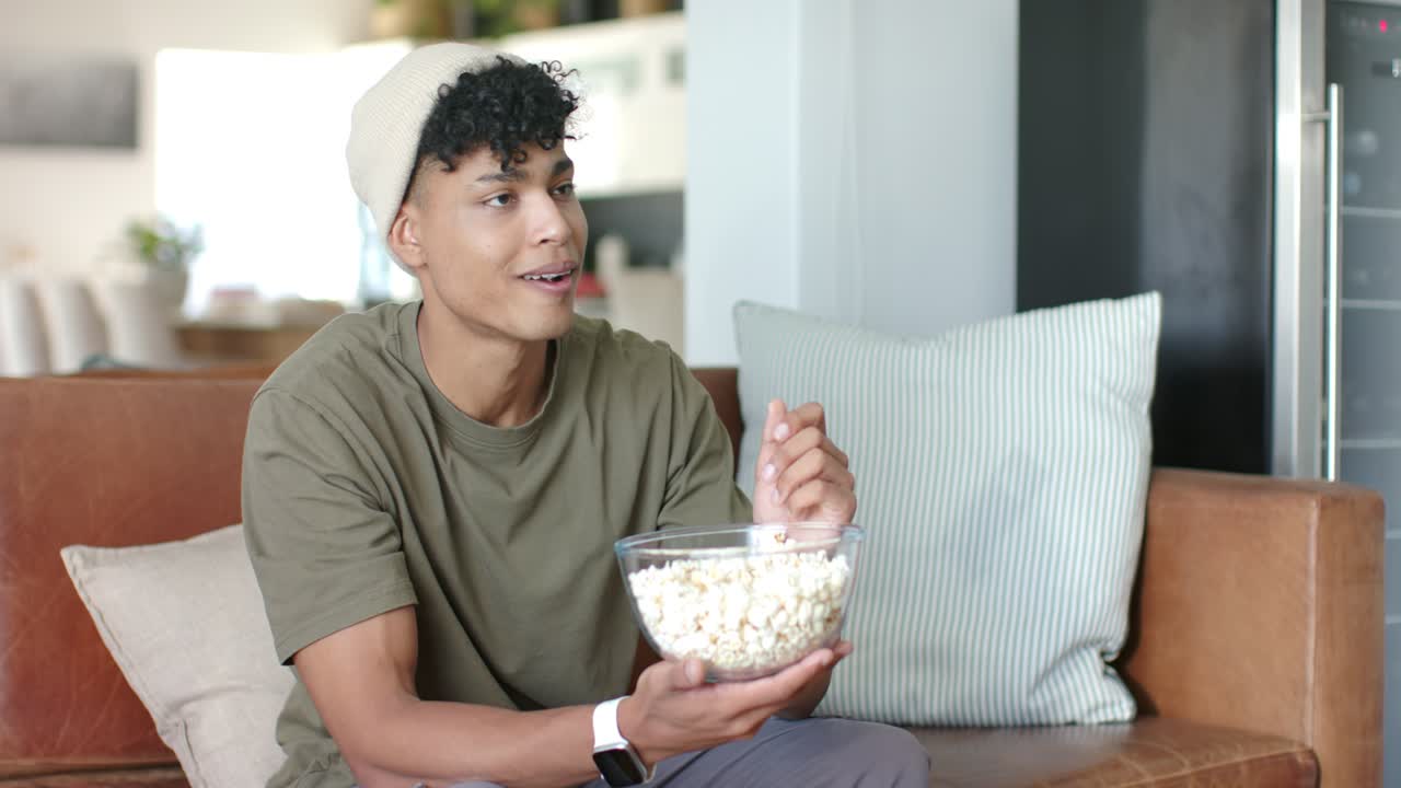 Uomo afroamericano che tiene in braccio una ciotola di vetro di popcorn sul divano, mangiando mentre reagisce a una scena fuori campo.