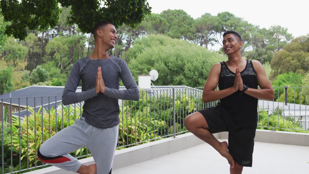 Smiling mixed race gay male couple standing on terrace practicing yoga