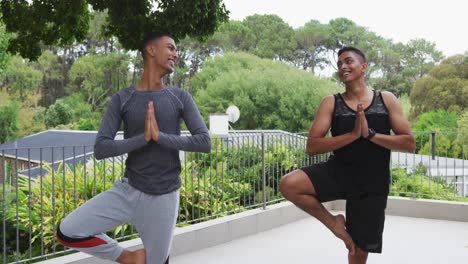Smiling-mixed-race-gay-male-couple-standing-on-terrace-practicing-yoga