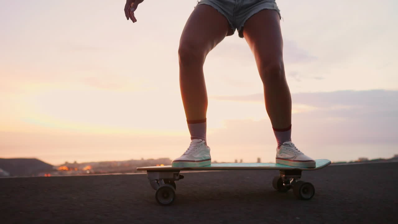 Slow motion captures the elegance of a beautiful young skateboarder riding her board in shorts along a mountain road during sunset, while the mountains provide a stunning backdrop