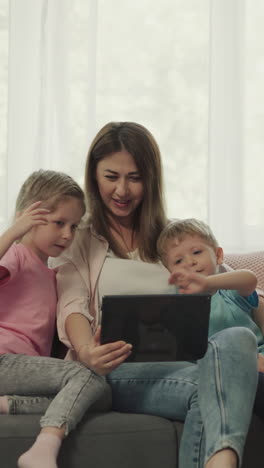 mother and children talk to father on video call via tablet waving hello with hands. happy family enjoys online communication sitting on sofa