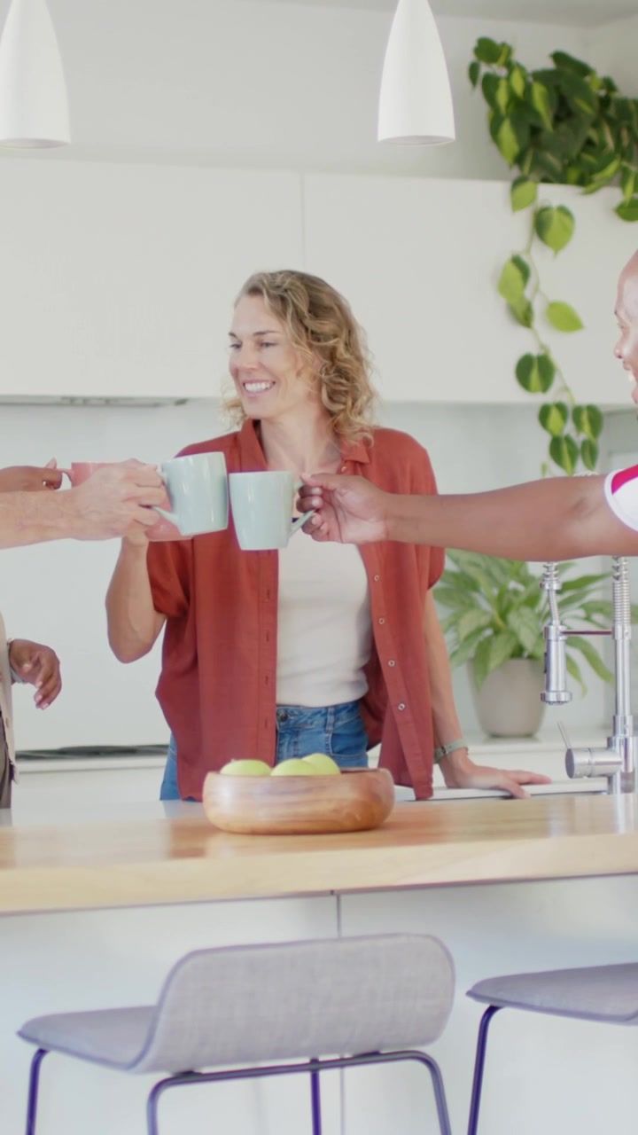 Mujer apoyada en la isla de la cocina, alcanzando una taza rosa mientras las manos se unen, chocando las tazas, celebrando un negocio.