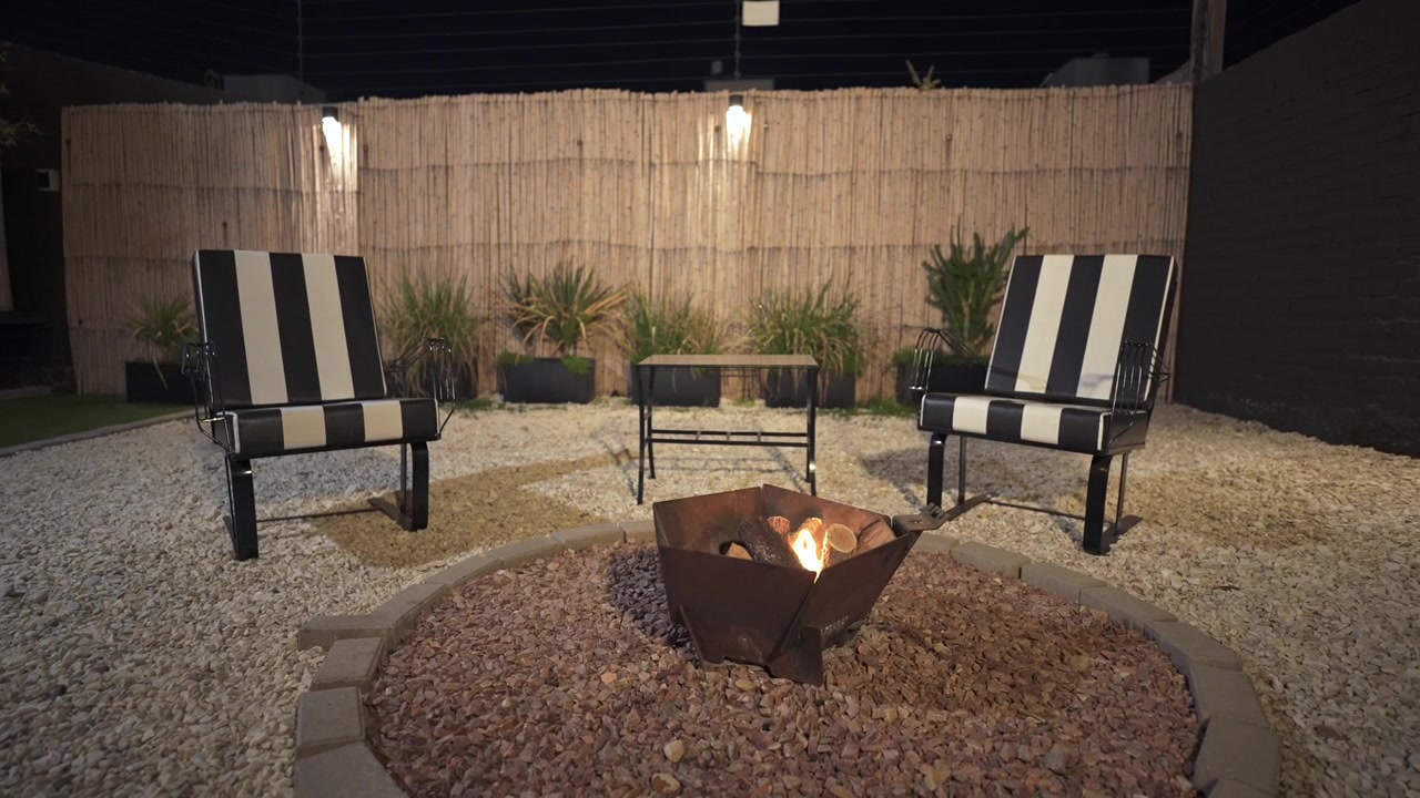 Una rilassante vista notturna di un moderno patio sul retro. Un fuoco caldo brucia in un braciere metallico circondato da ghiaia, sedie a righe bianche e nere e una recinzione di bambù illuminata.