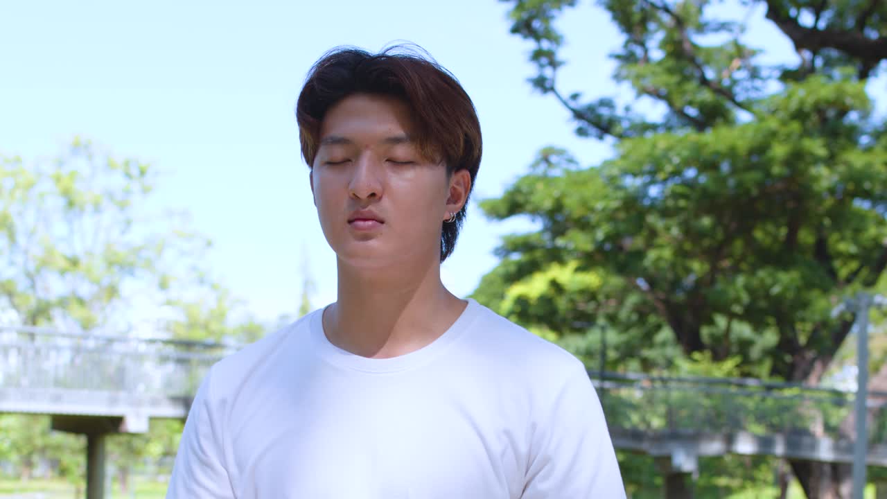晴れた都市公園で、若いアジア人男性が屋外でマインドフルネス瞑想をしている。