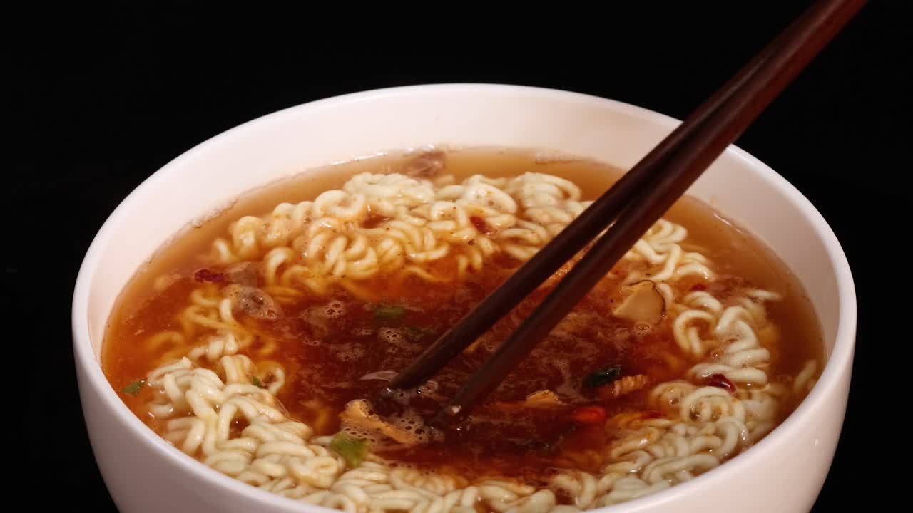 Un paio di bacchette mescola una ciotola di noodles istantanei con condimento in una ciotola bianca, sotto una luminosa illuminazione da studio su uno sfondo nero, ripreso in primo piano