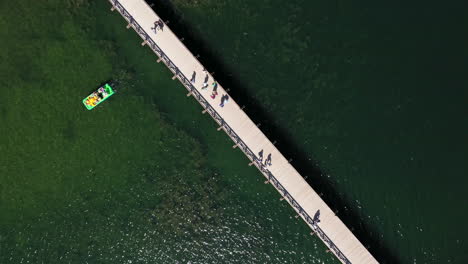 AERIAL:-Rotating-Shot-in-High-Altitude-of-Wooden-Bridge-in-Trakai-and-People-Crossing-It-with-Visible-Green-Color-Lake