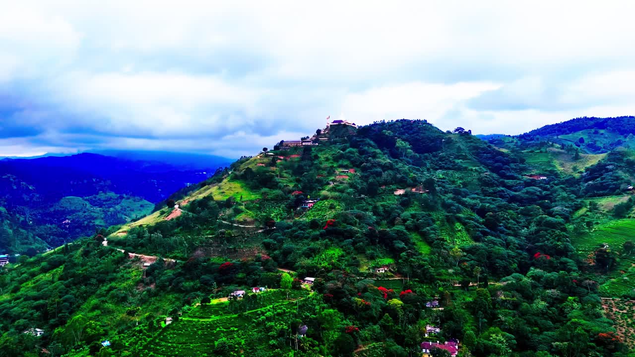 A scenic Sri Lankan mountain rises with a hilltop building, surrounded by green slopes, scattered village homes, tropical vegetation, and expansive mountain views.