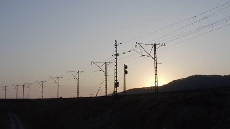 silueta de ferrocarril vacío con líneas eléctricas de alto voltaje y sol escondido detrás de una colina, al atardecer