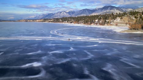 A-drone-passes-over-the-heads-of-a-couple-sitting-on-a-bench-on-top-of-the-frozen-surface-of-Lake-Invermere,-British-Columbia-Canada-with-skaters-skating-by