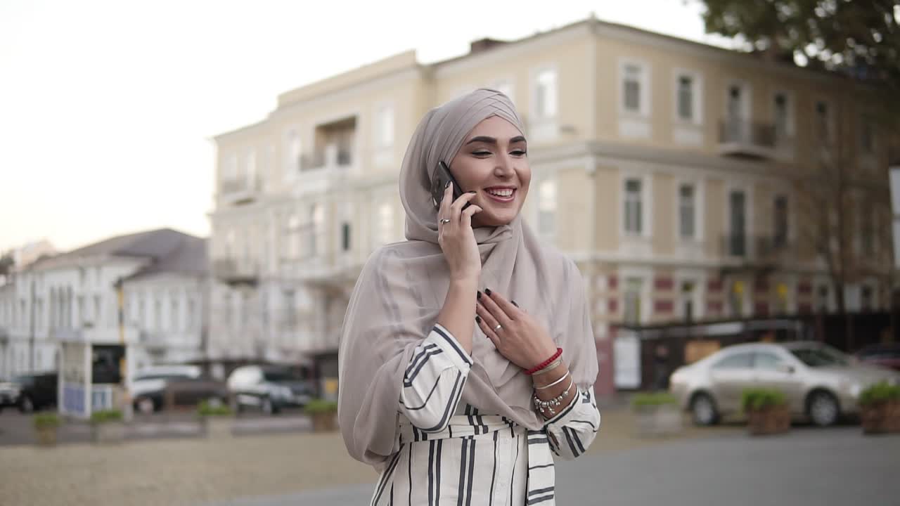 muslimsk kvinde i hijab med makeup, der går på gaden og taler i telefonen på baggrund af en smuk gammel klassisk bygning