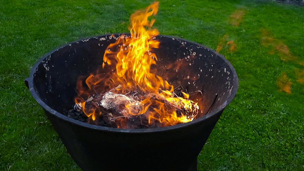 Bright orange flames and smoke rising from a black metal incinerator during a controlled burn in a backyard. The fire pit is sitting on a vibrant green grass lawn during the evening