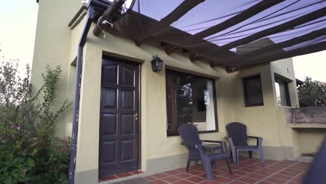 establishing shot of cozy outdoor patio featuring two chairs under a sunshade, attached to a modern home with a black door and large windows