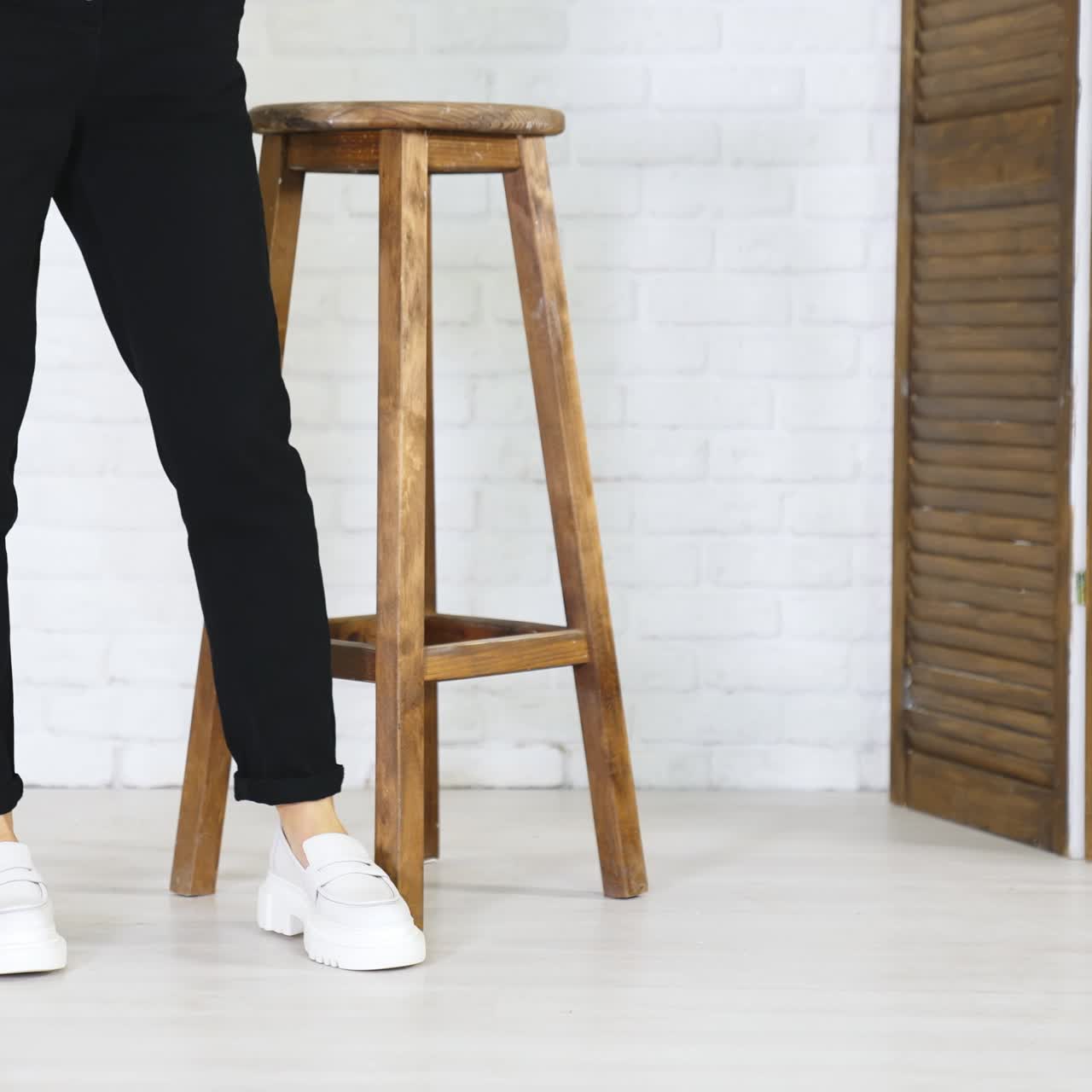 Girl model climbs up on the high wooden stool and puts her shoes on the footrest. White new modern footwear demonstration. White wall background