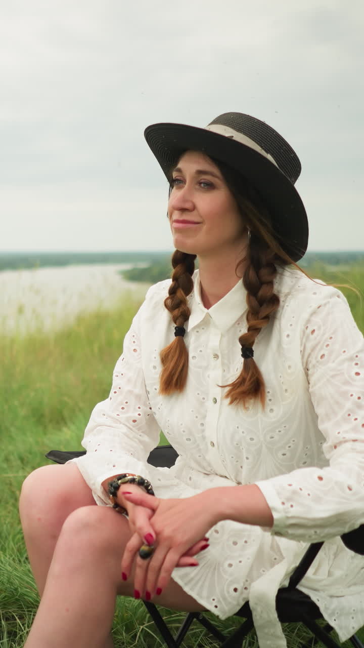 A beautiful woman wearing a hat and a white dress is seated on a chair in a lush grass field beside a tranquil lake, posing elegantly with a serene expression, under a cloudy sky