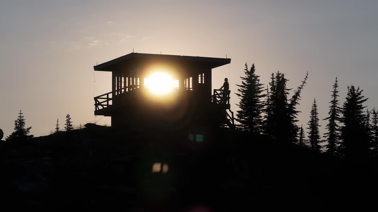 En panorering med drone fra luften, der bevæger sig fra højre mod venstre, viser et silhuet af et fjerntliggende, isoleret brandudkigstårn i det afsidesliggende nordlige Idaho, mens solen skinner gennem vinduerne, og en kvinde læner sig op ad et gelænder.