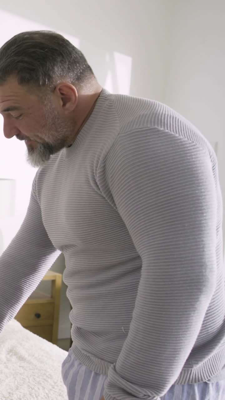 Video verticale: un uomo barbuto alza la mano, controlla un tablet accanto al letto, mettendosi seduto, con una maglia a costine.