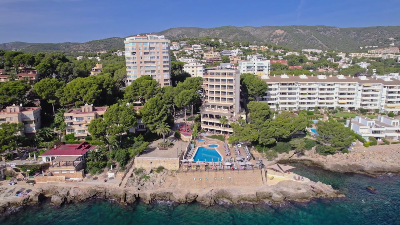 Um drone orbita lentamente um hotel resort em uma costa rochosa, revelando um terraço de piscina na falésia e colinas cobertas de pinheiros. Água turquesa encontra rocha acidentada sob um céu azul claro