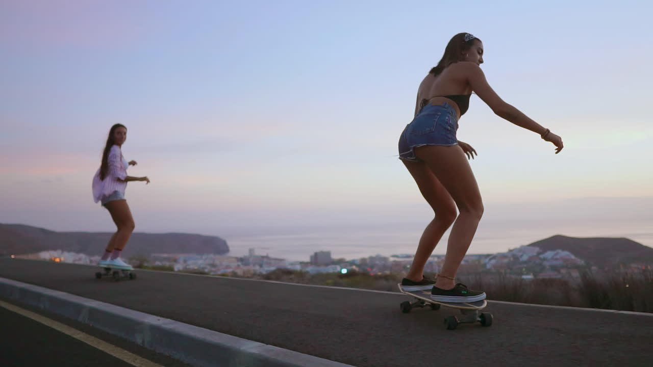 On a road at sunset, two friends skateboard in slow motion, with mountains and a scenic sky forming the backdrop. They're wearing shorts