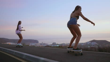 On-a-road-at-sunset,-two-friends-skateboard-in-slow-motion,-with-mountains-and-a-scenic-sky-forming-the-backdrop.-They're-wearing-shorts