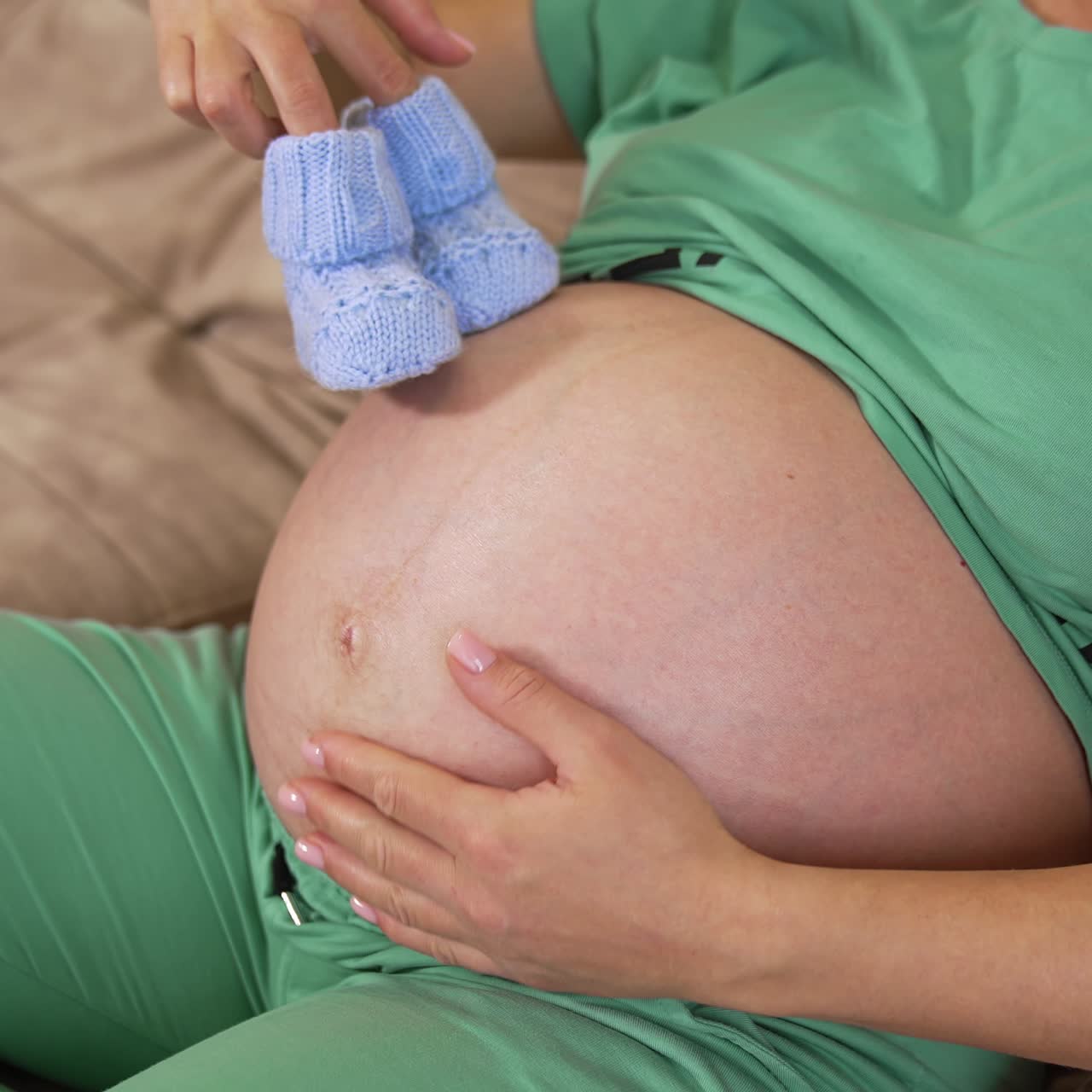 Giovane futura mamma che tiene in mano delle scarpine fatte a maglia. Donna incinta con un grosso ventre scoperto che gioca con dei calzini blu a casa. Gravidanza
