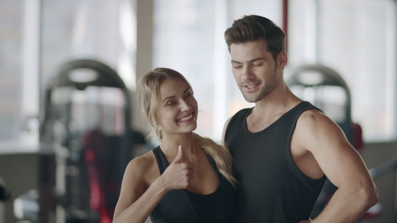 Casal feliz posando em academia moderna. Mulher sorridente fazendo sinal de positivo.
