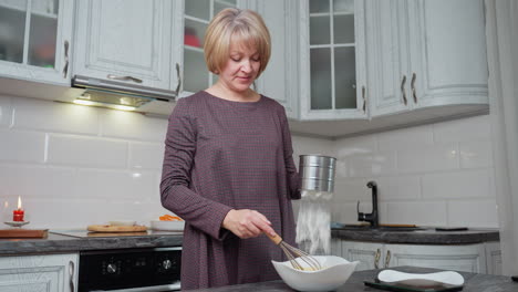 mujer agitando la mezcla en un cuenco blanco mientras tamiza la harina de trigo con un tamiz de acero inoxidable en la cocina moderna, preparación de horneado con configuración de encimera
