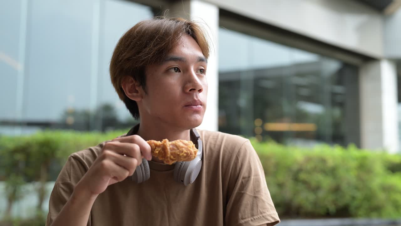 Young handsome Asian man eating fried chicken outdoors in restaurant during summer while sitting alone wearing casual t-shirt and headphones around neck
