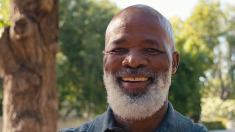 Close-Up-Portrait-Of-Smiling-Senior-Man-Standing-Outdoors-In-Garden-Park-Or-Countryside