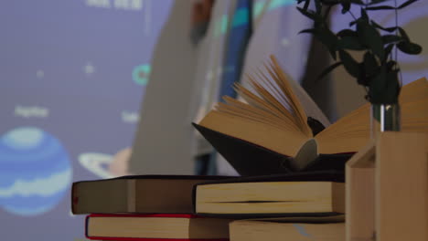 primer plano de los libros de texto en el escritorio mientras el maestro da una lección de matemáticas o ciencias en la escuela de pie en la parte delantera de la clase con la proyección de un gráfico