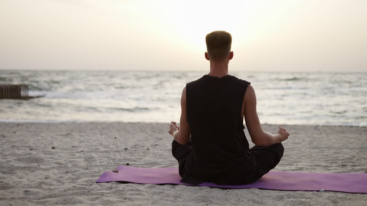 un joven está haciendo yoga y meditando en una alfombra deportiva mientras está sentado durante el amanecer del hijo. vista de atrás. haciendo un específico